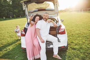 vovô e avó sentam-se juntos na natureza perto do carro. eles fazem uma selfie e a neta brinca com eles foto