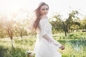 uma jovem com um vestido longo branco está caminhando no jardim. lindo pôr do sol através das folhas das árvores foto