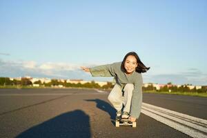 skatista menina equitação em skate, em pé em dela Longboard e rindo, equitação cruzador em a esvaziar rua para a Sol foto