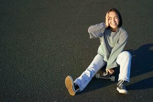 lindo coreano adolescente menina senta em skate e goza ensolarado dia. sorridente jovem ásia mulher patinação em Longboard foto