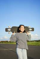 vertical tiro do despreocupado ásia menina com Longboard. jovem mulher skatista segurando cruzador em dela ombros e caminhando em estrada, skate foto