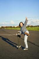 vertical tiro do feliz ásia skatista garota, pulando, em pé com skate e sorridente. mulher patinação em Longboard e tendo Diversão foto