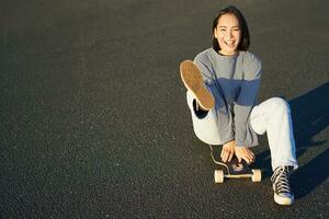 retrato do lindo ásia menina patinação, sentado em dela skate e sorridente. fofa adolescente com Longboard, patinação em estrada foto