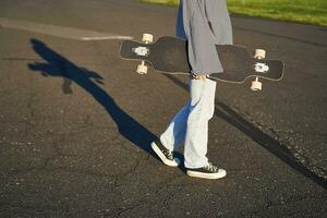 cortada tiro do adolescente menina corpo, segurando cruzador Longboard dentro mão, caminhando dentro tênis em estrada dentro jeans e suéter. jovem mulher skatista com skate foto