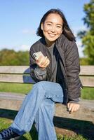 fofa jovem mulher com Smartphone dentro mãos, sentado em Banco e sorridente, usando Móvel telefone, esperando para alguém dentro parque foto