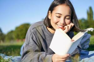 retrato do lindo sorridente ásia garota, lendo dentro parque, deitado em Relva com favorito livro. lazer e pessoas conceito foto