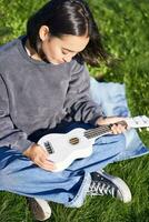 vertical tiro do menina músico, olhando com Cuidado às dela branco ukulele guitarra, jogando música dentro parque, sentado em Relva foto