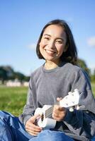 retrato do fofa sorridente menina jogando ukulele dentro parque. jovem mulher com musical instrumento sentado ao ar livre foto