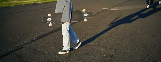 cortada tiro do adolescente menina corpo, segurando cruzador Longboard dentro mão, caminhando dentro tênis em estrada dentro jeans e suéter. jovem mulher skatista com skate foto