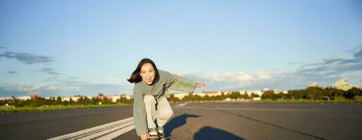 skatista menina equitação em skate, em pé em dela Longboard e rindo, equitação cruzador em a esvaziar rua para a Sol foto