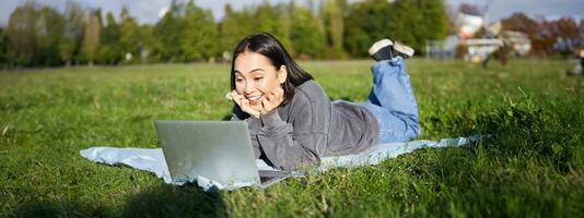 retrato do lindo menina deitado dentro parque e olhando com surpreso face às dela computador portátil, assistindo vídeos, conversando com amigos enquanto relaxante ao ar livre foto