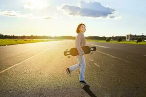 ásia menina com skate em pé em estrada durante pôr do sol. skatista posando com dela grandes quadro, cruzador área coberta durante Treinamento foto
