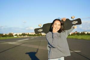 lazer e pessoas. feliz ásia mulher em pé com Longboard, cruzeiro em a esvaziar estrada dentro interior. skatista menina detém dela skate e sorrisos às Câmera foto