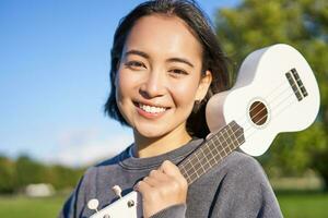 retrato do lindo sorridente menina com cavaquinho, ásia mulher com musical instrumento posando ao ar livre dentro verde parque foto