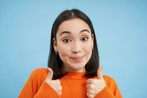 fechar acima retrato do ásia mulher mostra polegares acima, sorrisos e parece solidário, aprova, recomenda smth, dá positivo responder, azul fundo foto