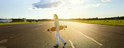 jovem skatista garota, adolescente patinação em cruzador, segurando Longboard e caminhando em concreto esvaziar estrada foto