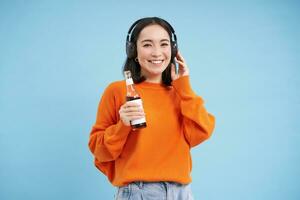 lindo ásia mulher escuta música dentro fones de ouvido, bebidas Coca a partir de garrafa e sorrisos às Câmera, azul fundo foto