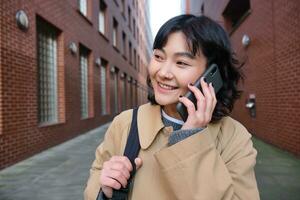 retrato do jovem coreano mulher caminhando baixa rua com telefone, falando com alguém, faz uma chamar, tem Telefone conversação foto