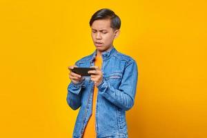 Homem asiático usando o celular, jogando o jogo com a cara zangada sobre o fundo amarelo foto