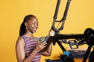 jovem Preto senhora inspecionando estragado bicicleta e explorando reparar opções em dela inteligente tábua. imagem do africano americano mulher verificação quebrado bicicleta componentes enquanto segurando uma digital dispositivo. foto