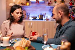 feliz homem surpreendente namorada com diamante luxo noivado anel desfrutando Natal feriado juntos sentado às jantar mesa dentro natal decorado cozinha. casado casal desfrutando inverno estação foto