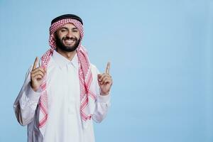 islâmico homem vestindo tradicional vestuário apontando para cima com dedos estúdio retrato. muçulmano pessoa exibindo acima às propaganda e olhando às Câmera com sorridente expressão foto