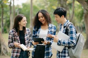 três jovem Faculdade alunos é lendo uma livro enquanto relaxante sentado em Relva dentro uma campus parque com dela amigos. Educação conceito foto