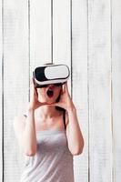 garota de óculos VR, animada, segurando as mãos no rosto, fundo de madeira branco foto