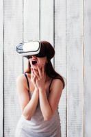 linda garota de óculos VR, animada, segurando as mãos no rosto, fundo de madeira branco foto