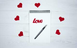 em branco caderno, caneta, vermelho corações e a palavra amor em uma branco de madeira fundo. romântico cumprimento para dia dos namorados dia. topo visualizar. foto