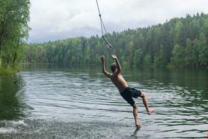 Garoto saltos para dentro a água usando uma Tarzan balanço enquanto natação dentro uma floresta lago foto