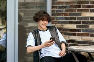 jovem homem lendo engraçado mensagem em telefone enquanto sentado às a ao ar livre cantina mesa foto