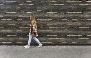 jovem mulher anda em baixa a rua contra uma tijolo parede foto