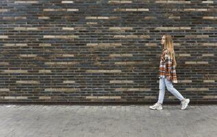 jovem mulher anda em baixa a rua contra uma tijolo parede foto