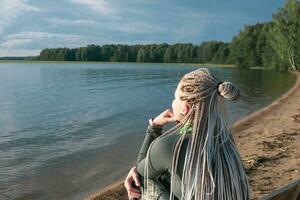 mulher viajante com de tranças Penteado sentado em uma arenoso costa do uma grande lago foto