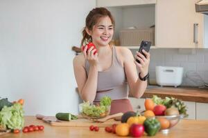 lindo jovem mulher dentro exercício roupas tendo Diversão dentro uma fofa cozinha às lar. usando a telefone para estude em formação e preparar vegano fruta salada curativos, fruta treme, ou saudável batidos. foto