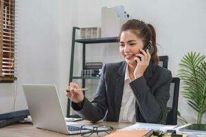 empresária falando em o negócio telefone e usando computador portátil para Faz finança, matemática em de madeira mesa dentro escritório e o negócio fundo, imposto, contabilidade, Estatisticas e analítico pesquisa conceitos. foto