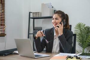 empresária falando em o negócio telefone e usando computador portátil para Faz finança, matemática em de madeira mesa dentro escritório e o negócio fundo, imposto, contabilidade, Estatisticas e analítico pesquisa conceitos. foto