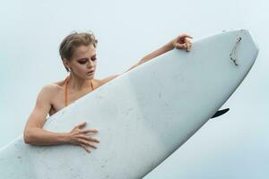 fêmea Esportes moda modelo segurando prancha de surfe. ela é em pé atrás surfar borda e olhando baixa foto