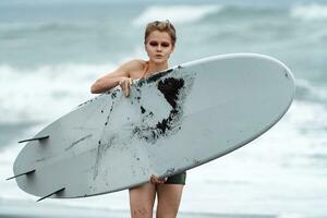 mulher surfista carregando branco prancha de surfe caminhando em praia, olhando às Câmera em fundo do mar ondas foto