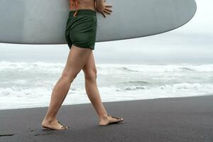 surfista mulher carregando prancha de surfe caminhando ao longo de praia. lado Visão do fêmea pernas e nádegas foto