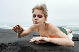 retrato Esportes moda modelo com brilhante Maquiagem e fechadas olhos deitado em prancha de surfe em arenoso de praia foto