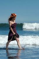 ruiva mulher é desfrutando verão feriados de caminhando tornozelo profundo dentro refrescante oceano ondas em de praia foto
