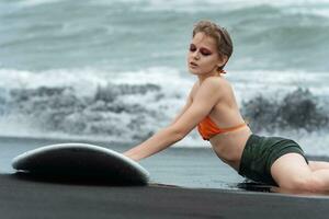 muito quente sexy mulher surfista é deitado em Preto areia do de praia com prancha de surfe e olhando baixa foto