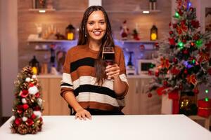 retrato do alegre mulher segurando vidro do vinho dentro festivo cozinha decorado para Natal véspera jantar celebração. caucasiano jovem pessoa desfrutando beber dentro inverno estação às casa foto