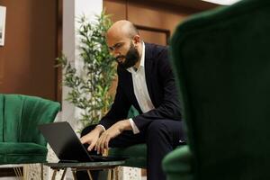 homem de negocios usando computador portátil às hotel, procurando para soluções para presente às internacional reunião. branco colarinho trabalhador esperando para começar conferência dentro salão área, forjamento Novo colaborações. foto