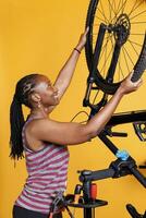 enérgico africano americano fêmea desmantelamento estragado bicicleta pneu para reparar e substituir com novo. jovem Atlético Preto mulher ganancioso e carregando bicicleta roda para mais distante manutenção. foto