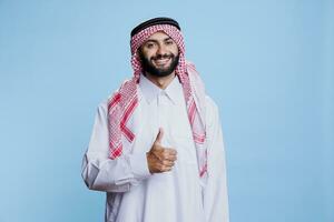 alegre muçulmano homem vestindo tradicional islâmico roupas mostrando polegar acima retrato. sorridente árabe pessoa em pé com aprovar placa e olhando às Câmera com despreocupado expressão foto