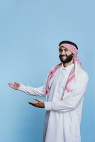 animado árabe vendas pessoa vestido dentro tradicional islâmico roupas publicidade produtos, apontando para lado com mãos. alegre muçulmano homem mostrando cópia de espaço e olhando às Câmera foto