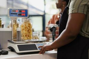 detalhado tiro do vendedor utilizando digital escala para determinar peso do recipiente preenchidas com orgânico produzir. fechar-se do fornecedor pesagem jarra do fresco massa, com cliente pacientemente esperando às contador. foto